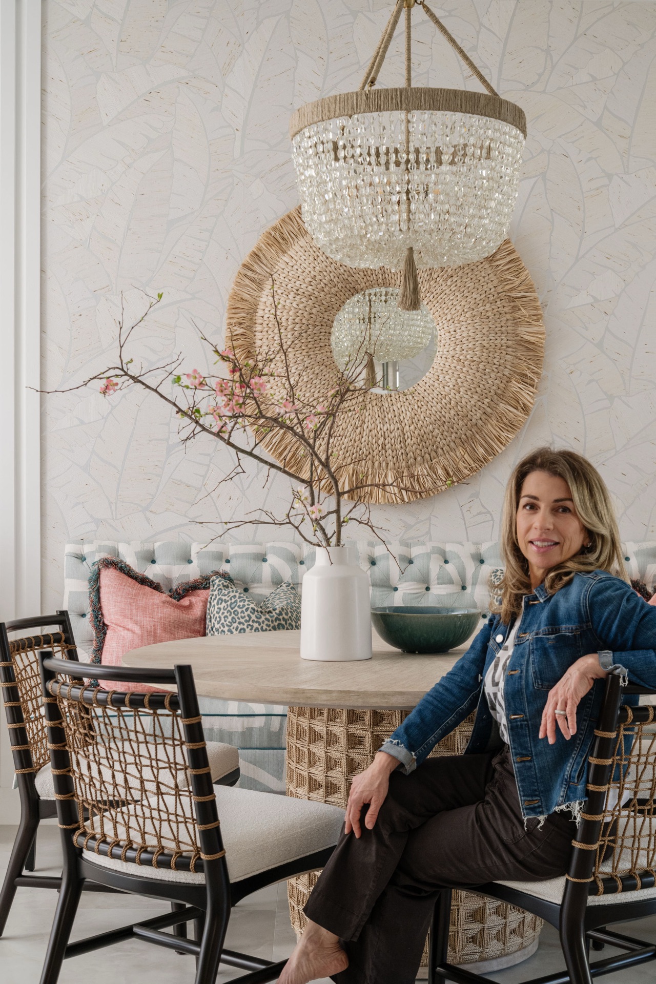 Nina Asadoorian, principal designer and founder of Oak Heart Interiors, seated in a softly layered dining space with a woven mirror and crystal chandelier